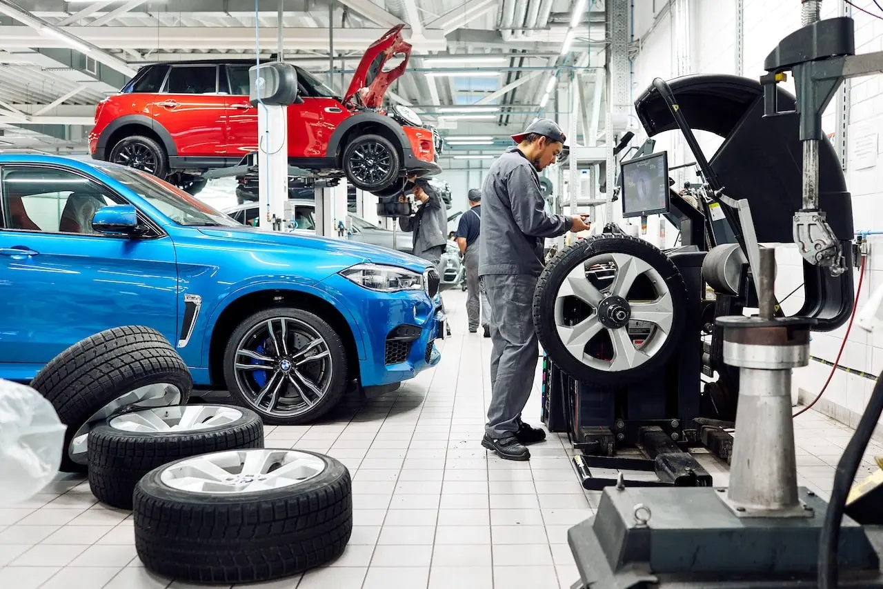 Mechanics in a garage work on cars and tire alignment. A blue car and a red SUV are visible, with tires and equipment scattered around the well-lit workspace.